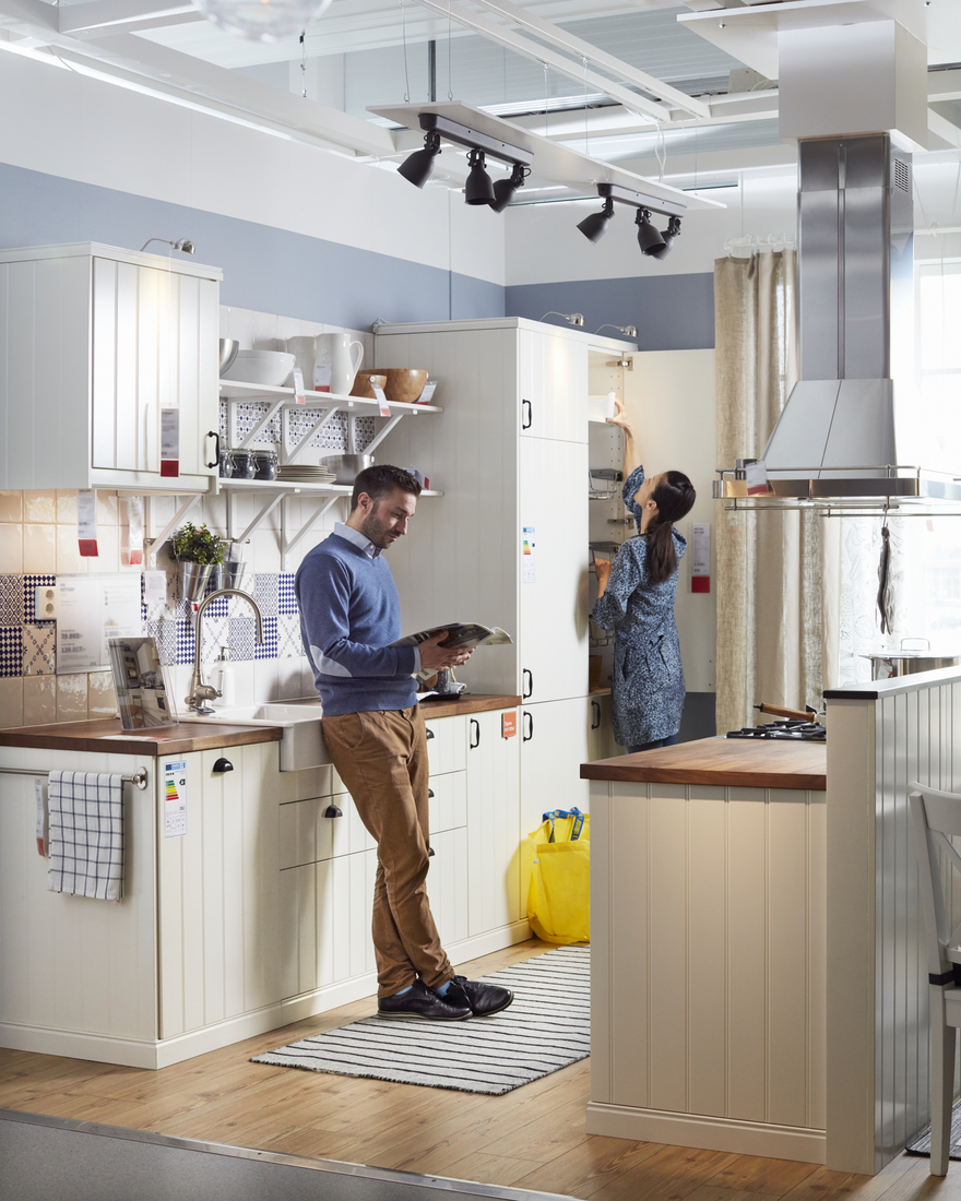Man reading a catalog in a showroom kitchen while a woman checks an upper cabinet.