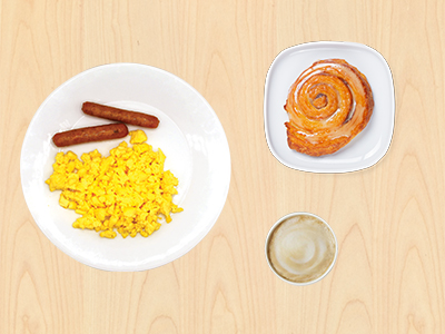 Vista superior de un desayuno sobre una mesa de madera clara, que incluye un plato de huevos revueltos con dos salchichas, un plato separado con un rollo de canela y una taza de café.