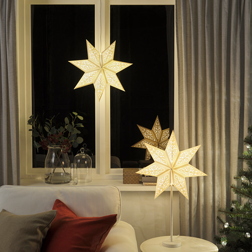 Lámpara STRÅLA: pantalla de cartón blanco en forma de estrella. Resplandor pequeño, brillante y cálido. En la mesa cerca de la ventana, el sofá y el árbol de Navidad.