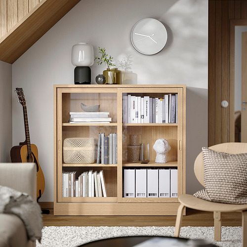 Moderna librería TONSTAD de roble claro con puertas correderas, que contiene libros y decoración, enmarcada por un reloj blanco y plantas, junto a una guitarra.
