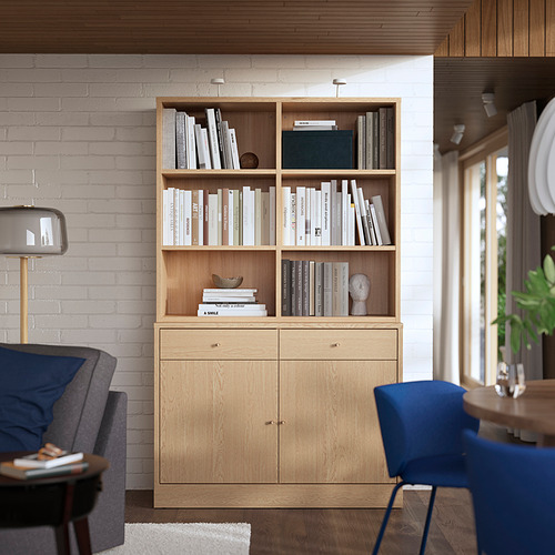 Estantería de madera con almacenamiento abierto y cerrado, llena de libros. Contra una pared de ladrillo blanco, junto a una silla gris y un taburete azul.