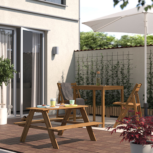 Pequeña mesa de picnic RESÖ de madera en la cubierta, rodeada de sillas a juego, cerca de la casa con una gran ventana, perfecta para los niños.