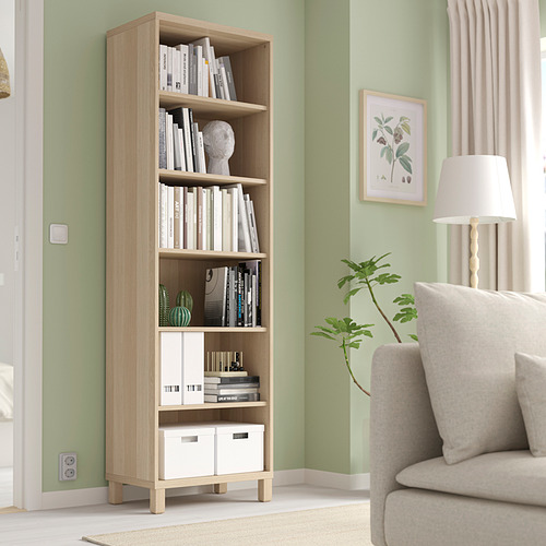 Estantería BESTÅ en roble claro con libros y decoración contra la pared verde. Sillón beige y lámpara cerca.