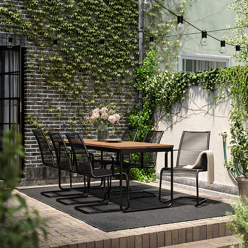 Moderno comedor al aire libre con mesa y sillas VÄSMAN de metal negro, rodeado de vegetación y guirnaldas de luces.