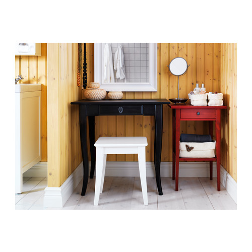 Un baño de madera bien organizado con escritorio negro, pequeño taburete blanco, mesa auxiliar roja, cestas tejidas y toallas enmarcadas sobre un piso de baldosas blancas.