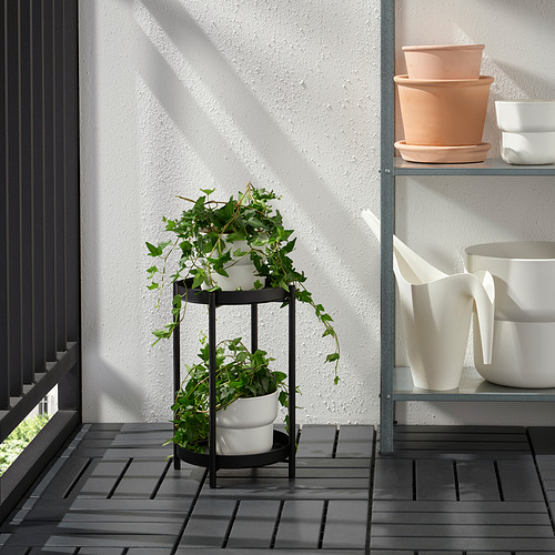 Soporte de plantas de metal de dos niveles con macetas blancas que sostienen plantas verdes en un balcón, junto a un estante de metal con macetas adicionales.