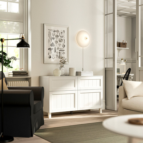 Salón con aparador blanco de BESTÅ , lámpara, estampado botánico y gran ventana. Sofá gris oscuro a la izquierda, objetos decorativos en el aparador.