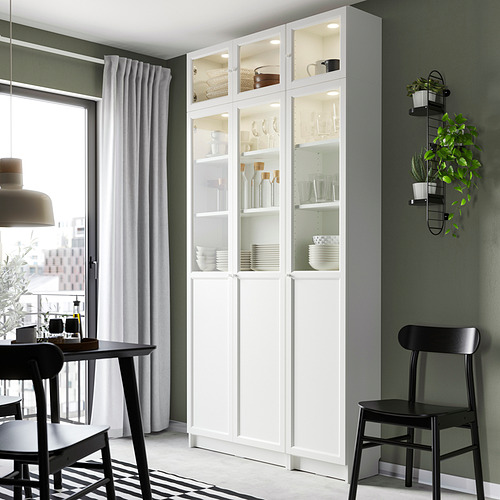 Moderno comedor con vitrina blanca BILLY que exhibe platos, flanqueada por sillas negras y un gran ventanal con cortina.