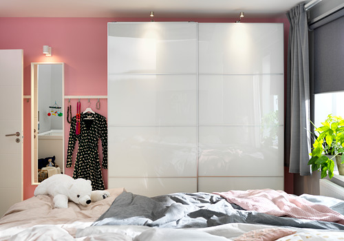 Dormitorio con armario de cristal blanco FÄRVIK, paredes rosas, cortinas grises y cama deshecha con un osito de peluche.