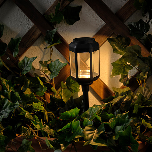 Una lámpara encendida con energía solar entre exuberantes plantas verdes, cuya cálida luz brilla a través de paneles transparentes, montados sobre una estructura de madera.
