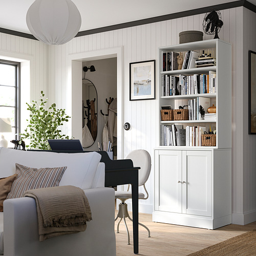 Una estantería blanca HAVSTA llena de libros y decoración, junto a un sofá gris con manta y un ordenador portátil.