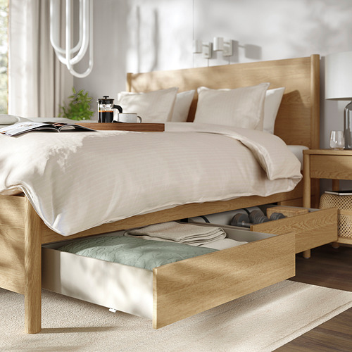 Cama moderna con estructura de madera, cajón abierto que muestra la ropa de cama, sábanas blancas en la cama, mesa auxiliar con configuración de café, planta y luz suave.