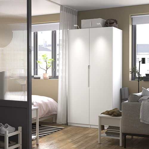 Dormitorio moderno con armario PAX blanco. Armario alto de dos puertas, encima de una mesita, cerca de una silla gris. Iluminación tenue, suelo de madera.