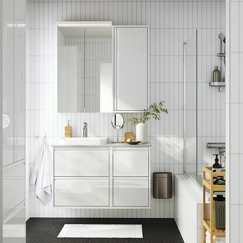 Moderno baño blanco con armario alto ÄNGSJÖN, lavabo, espejo, papelera negra, toallas, planta y carrito.