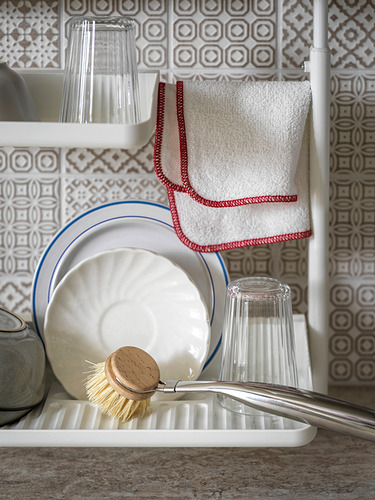 Estante de cocina SUNNERSTA blanco con platos, vidrio y cepillo. Pared de azulejos grises, toallas con bordes rojos.