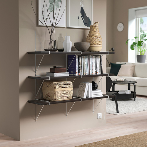La estantería de pared negra BERGSHULT, que contiene varios artículos, se mantiene alta, llenando un rincón junto a una pared beige, con un diseño moderno y minimalista.