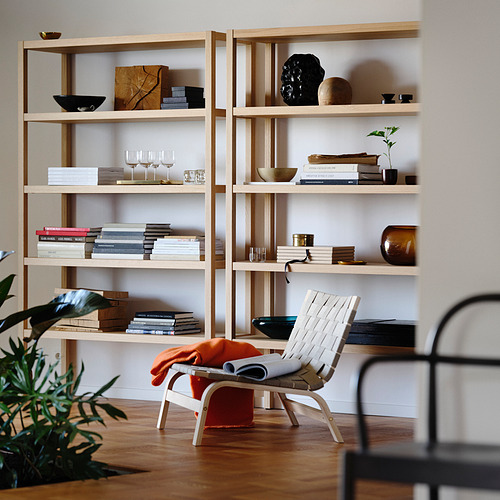 Sala de estar moderna con estanterías de madera clara, una silla blanca de estilo escandinavo con manta naranja y varios elementos decorativos.