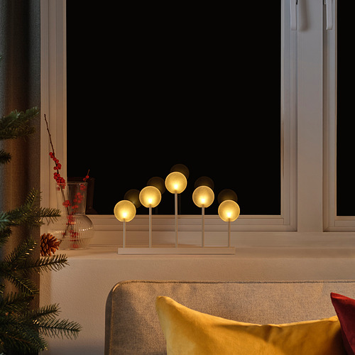 Acogedor candelabro STRÅLA de escena interior con cálidas luces led en el alféizar de una ventana, flanqueado por decoraciones festivas y una almohada amarilla.