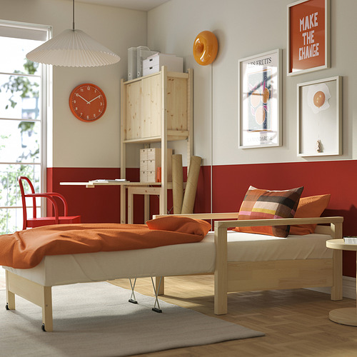 Dormitorio luminoso con cama TORNSBORG plegada como sillón, manta naranja, reloj rojo, arte y escritorio de madera.