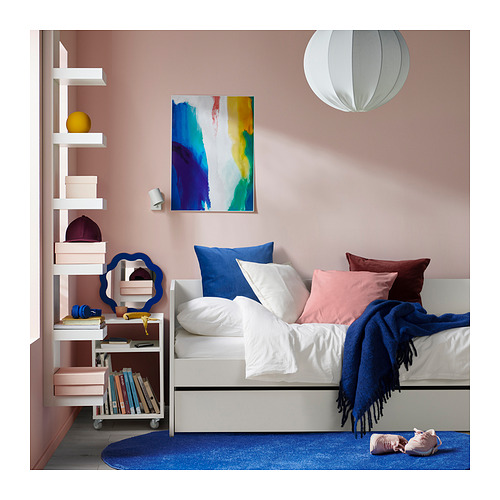 Acogedora habitación con sofá cama VIHALS blanco, almohadas de colores, alfombra azul, estantería con libros y arte abstracto en la pared rosa.