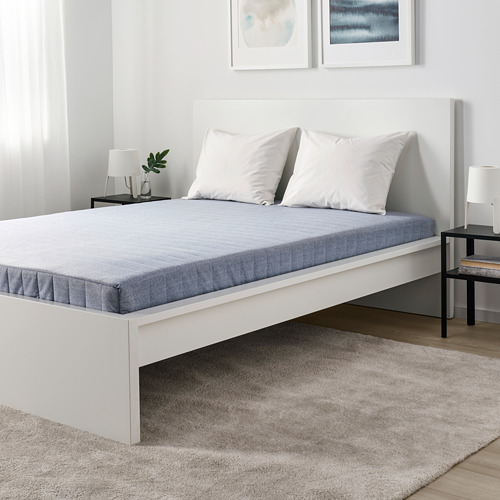 Cama minimalista blanca con colchón y almohadas grises, prominente y de perfil bajo en la decoración.