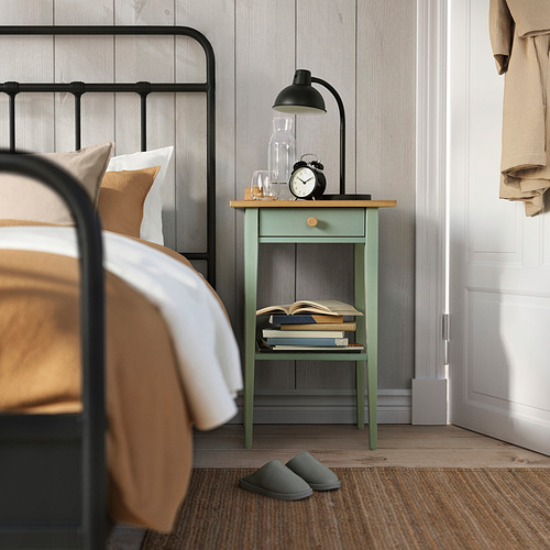 Acogedor dormitorio con mesita de noche HEMNES verde menta, lámpara de sostenimiento, reloj, libros, debajo de una estructura de cama de metal negro. Zapatillas debajo.