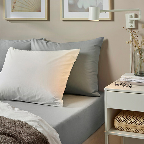 Acogedor dormitorio con ropa de cama de algodón DVALA gris, que incluye una sábana ajustable, dos almohadas y una manta. Mesita de noche decorativa con libros y jarrón.