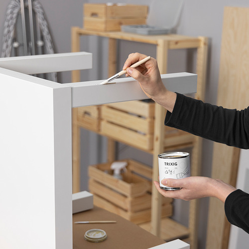 Persona pintando muebles blancos con un pincel usando lata de pintura TRIXIG. Estanterías de madera y herramientas al fondo.