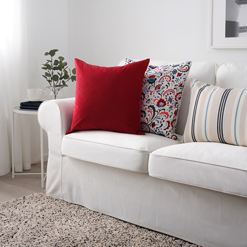 Sofá blanco con cojín de terciopelo rojo, almohadas florales y a rayas, planta y taza en la mesa auxiliar.