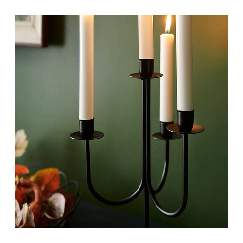Candelabro de metal negro con tres velas blancas. Se enciende una vela, que emite una luz suave. Colocado sobre mesa verde con flores.