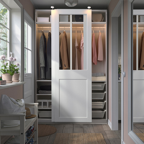 Un armario blanco PAX con puertas de cristal, exhibiendo ropa. Es grande, rodea una silla blanca más pequeña, e incluye cestas de malla para su almacenamiento.