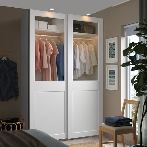 Armario blanco PAX con puertas de vidrio, luces, ropa, junto a ventana y silla.