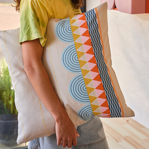 Persona sosteniendo un cojín OMMJÄNGE con motivos geométricos en la cama.