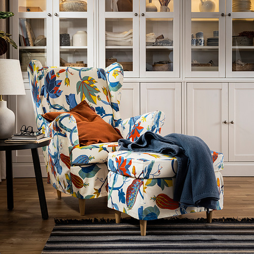 Silla floral STRANDMON con almohadas naranjas/azules, mesa auxiliar, lámpara, alfombra de rayas, armarios blancos.