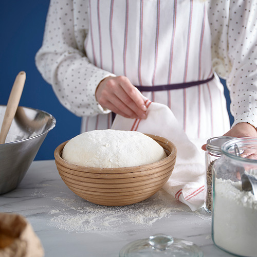 Una persona coloca la masa en una cesta redonda de madera espolvoreada con harina, rodeada de utensilios e ingredientes.
