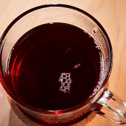 Taza de vidrio transparente llena de bebida oscura, burbujeante y especiada. Los vapores visibles sugieren calor. Descansando sobre una superficie de madera.