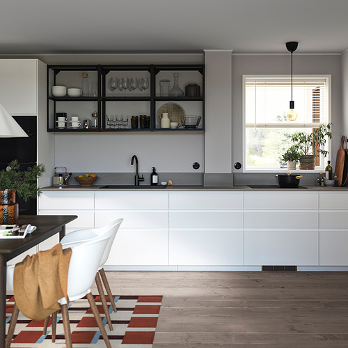Cocina moderna con gabinetes VOXTORP blancos, piso de madera y estantes abiertos con vasos y vajilla. También se ve una silla blanca y una alfombra de colores.
