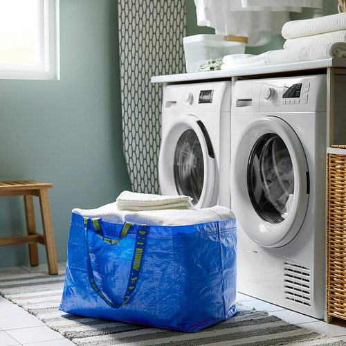 Un lavadero con una bolsa FRAKTA doblada en azul que contiene toallas. Dos lavadoras y toallas dobladas en la parte superior.