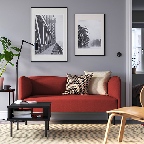 Sala de estar moderna con sofá LILLEHEM rojo, almohadas estampadas, mesa de centro negra, impresiones artísticas, lámpara de pie y planta en maceta.
