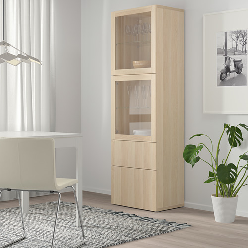 El mueble con puerta de cristal BESTÅ muestra cristalería y platos, cajones para almacenamiento, entre la mesa del comedor y la ventana con cortinas.