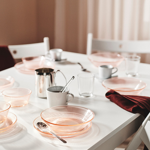 Vajilla de vidrio rosa sobre mesa blanca, con platos, tazones, tazas y cubiertos, que muestra un diseño elegante y durabilidad.