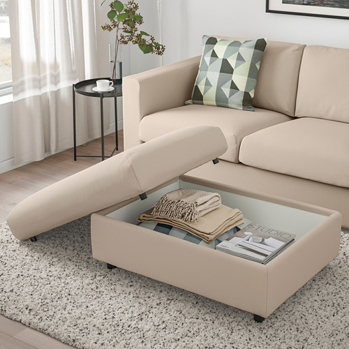 Sala de estar con sofá VIMLE beige, reposapiés con almacenamiento abierto que muestra mantas, revistas y ruedas. Mesa auxiliar con taza y planta.