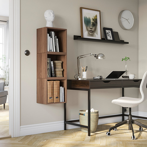 Oficina en casa con silla blanca, escritorio negro, estantes de madera, lámpara, computadora portátil, plantas. Las estanterías de EKET contienen libros, cestas. El reloj y el arte decoran la pared.