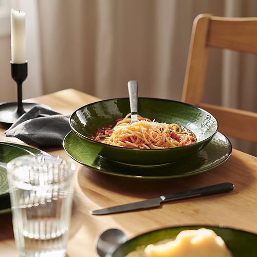 El cuenco de cerámica verde sostiene la pasta con un tenedor; colocado sobre una mesa de madera con vidrio, cuchillo y silla.