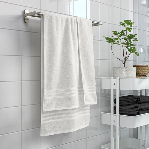 La toalla blanca BROKGLIM cuelga cuidadosamente, con bordes. Rodeado de toallas grises, plantas y azulejos. Percha de acero inoxidable.