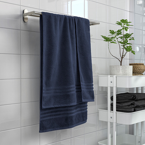 Toalla BROKGLIM azul sobre varilla de acero inoxidable, cerca de estantería blanca con toallas dobladas y planta.
