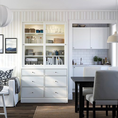 Unidad de almacenamiento HAVSTA blanca con puertas de vidrio, sofá, mesa de comedor y lámpara en una elegante sala de estar.