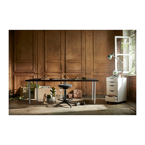 Habitación con paneles de madera con un moderno escritorio negro, taburete ajustable y gabinete blanco de 3 cajones. La iluminación natural de una ventana resalta la configuración.