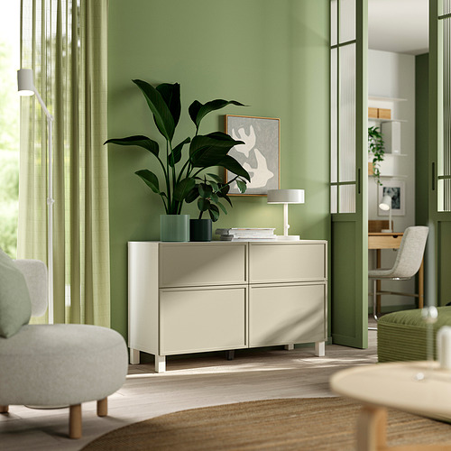 Sala de estar moderna con unidad de almacenamiento BESTÅ de color beige claro, planta de hojas grandes, arte abstracto, iluminación suave y área de comedor visible en el fondo.