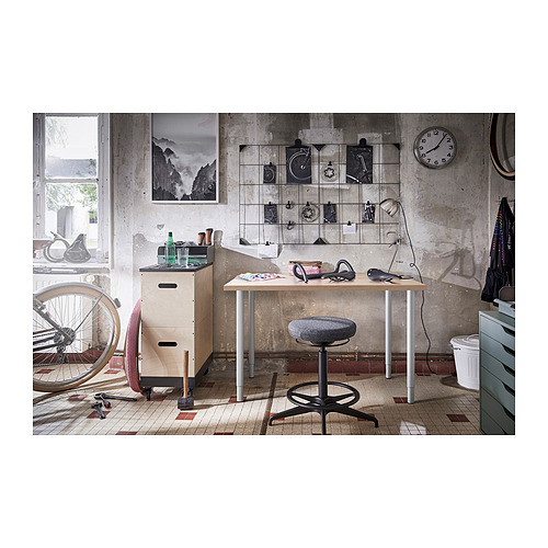 Oficina industrial con mesa LINNMON, reloj de pared, bicicleta y armario de almacenaje. Paredes de hormigón, suelo de baldosas y decoración variada.